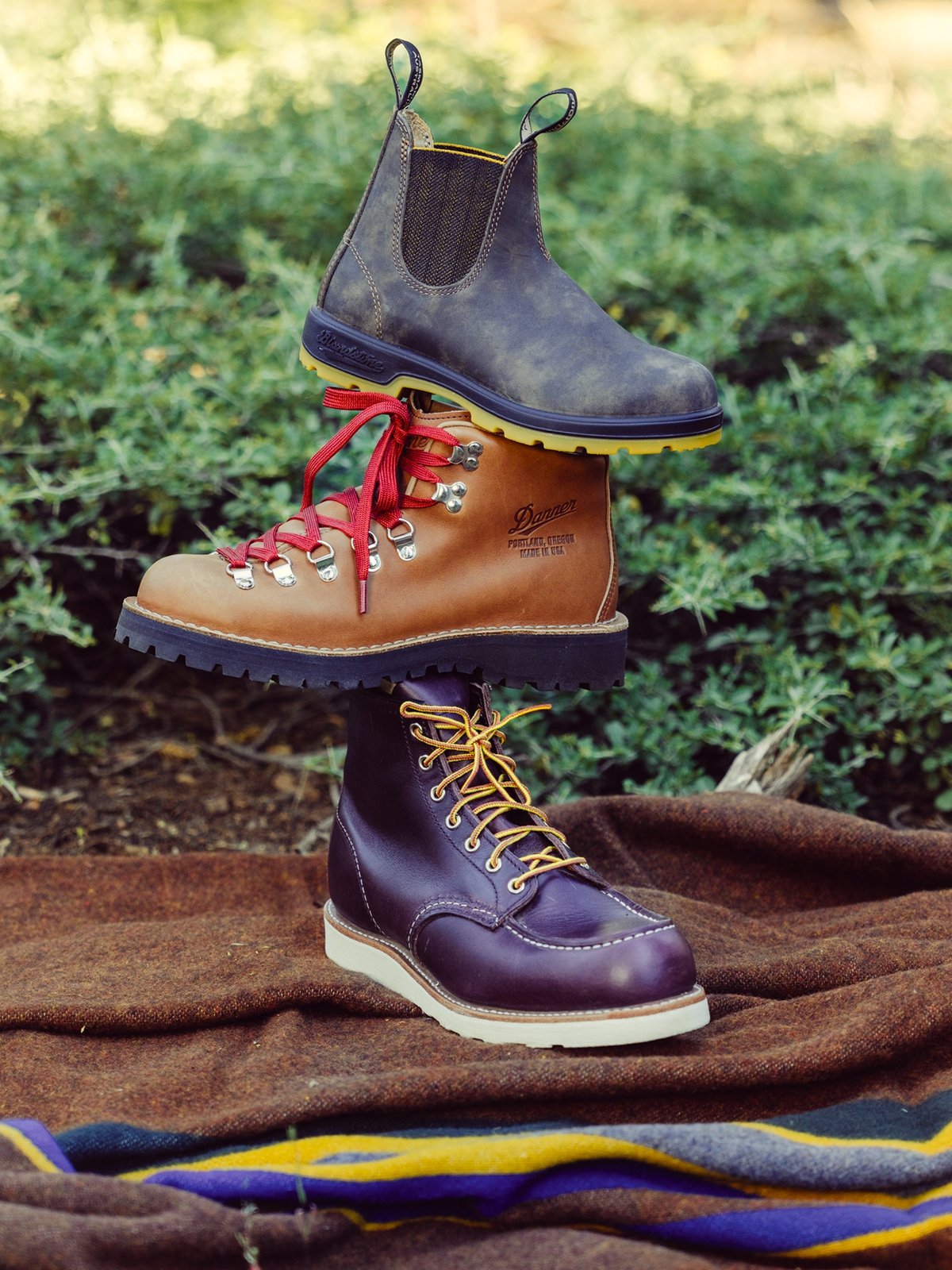 A Blundstone boot, a Danner hiking boot, and a Red Wing work boot are stacked on top of each other outdoors, all placed on a brown blanket.