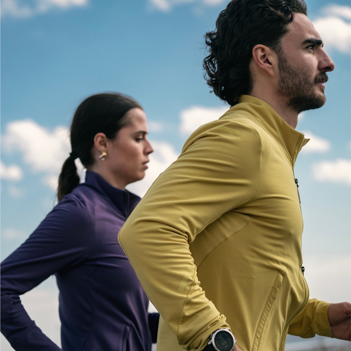A man in a yellow jacket and a woman in a purple jacket are running outdoors under a blue sky with clouds.