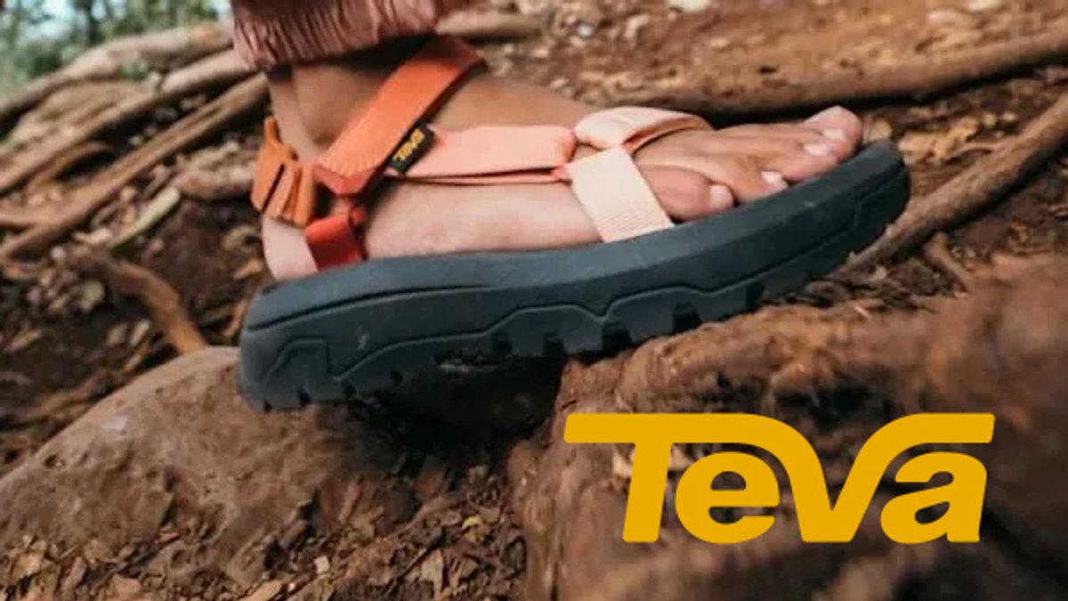 A person wearing orange Teva sandals with adjustable straps stands on rocky terrain.
