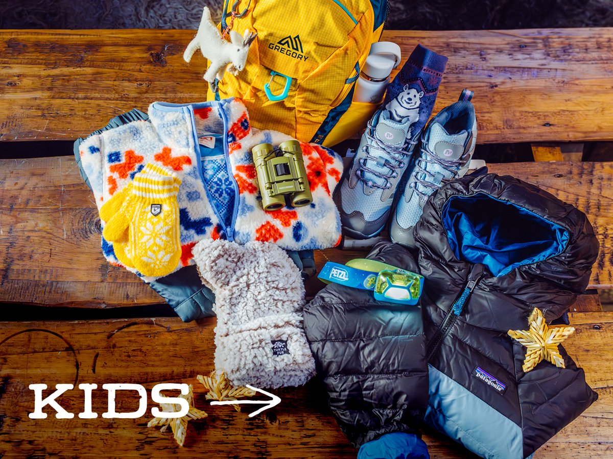 Kids’ outdoor gear, including a daypack, patterned fleece, mittens, binoculars, hiking shoes, a headlamp, and a puffy jacket, on a wooden table: “Kids.”