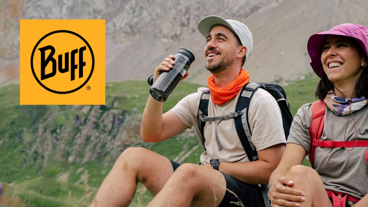 Two hikers wearing backpacks smile while taking a break on a mountainside, with one holding a Buff water bottle, showcasing outdoor adventure gear in a scenic valley landscape.