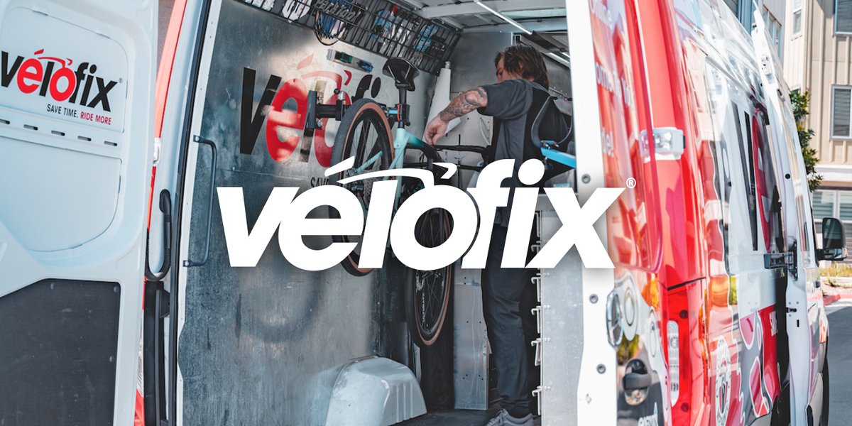 Mechanic working on a teal gravel bike mounted in a repair stand inside the open rear of a mobile service van. Featuring the Velofix logo.