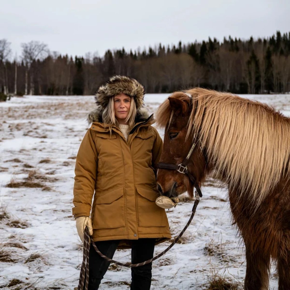 A woman wearing the Fjallraven Nuuk Parka stands against a snowy forest landscape. She holds reigns in her hand that lead to a nearby horse.