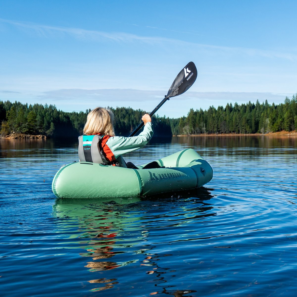 A person paddles a green inflatable kayak on a calm lake with a tree-lined shore in the background.