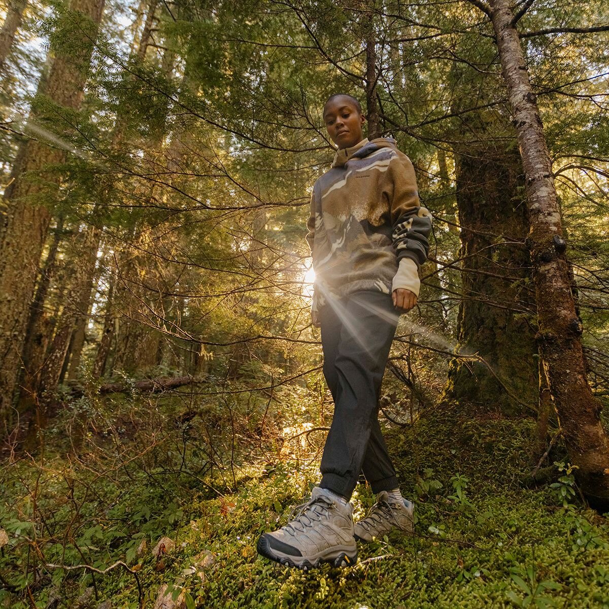 A woman walks through a sunlit Pacific Northwest forest wearing Merrell hiking shoes, dark pants, and a patterned hoodie.