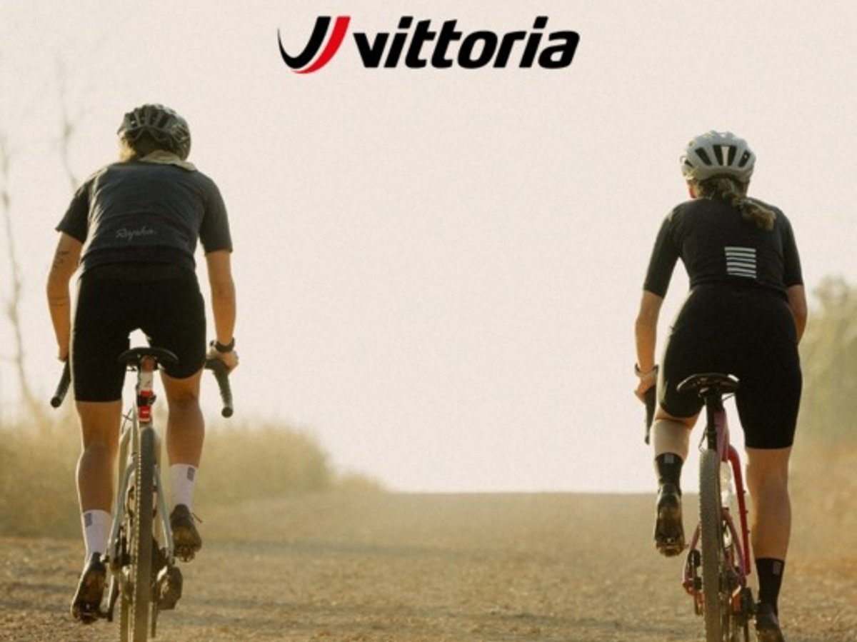 Two cyclists riding side-by-side down a dirt road toward the horizon; the Vittoria logo is centered at the top.