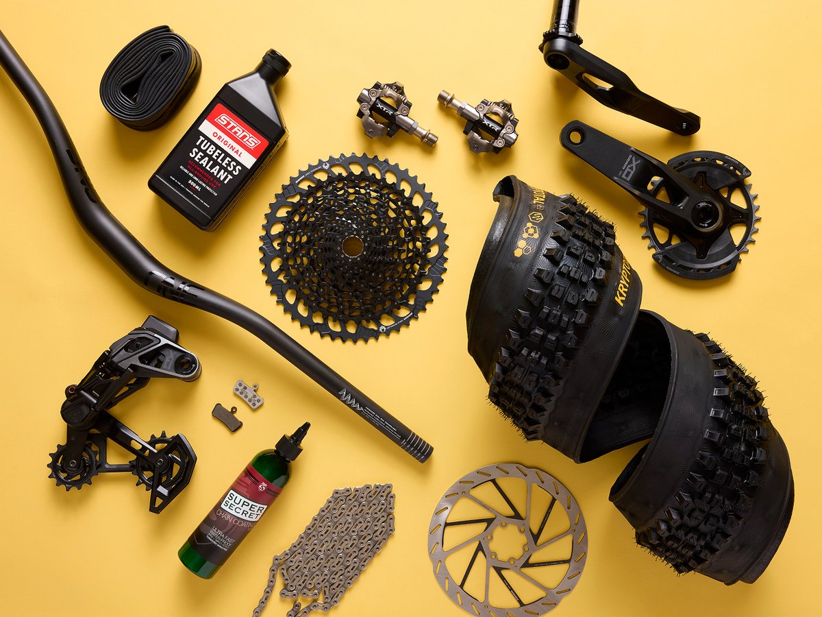 Flat-lay of mountain bike components — crankset, cassette, chain, derailleur, brake rotor, pedals, tubeless sealant, and knobby tires.
