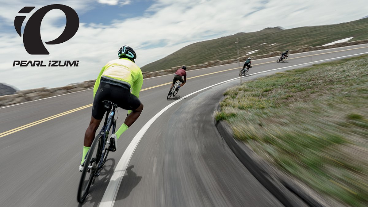 A cyclist in a high-visibility yellow jacket leads a group down a winding mountain highway, with the PEARL iZUMi logo in the corner.