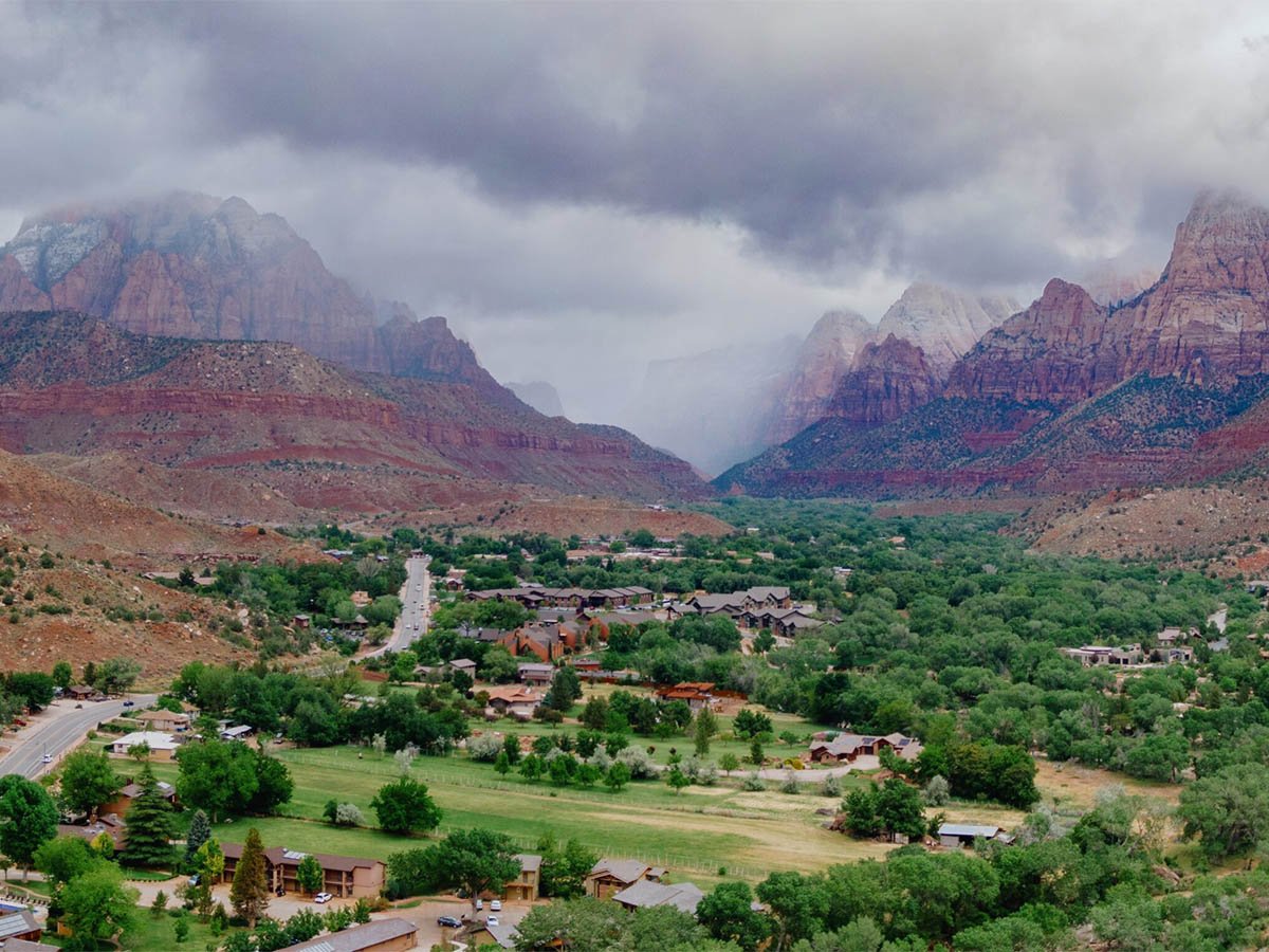 Aerial view of Springdale looking into Zion Canyon, with the town's tree-lined streets and lodges nestled between red sandstone peaks partially obscured by storm clouds.