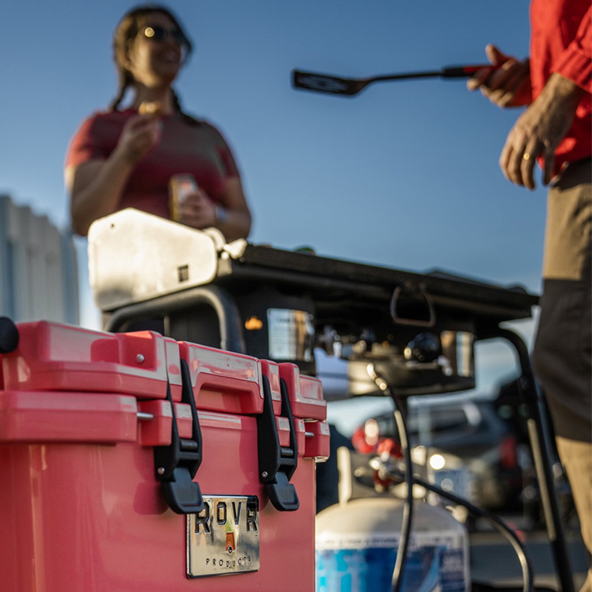 Coral RovR RollR cooler beside a portable grill at a tailgate, with people cooking and holding drinks in the background