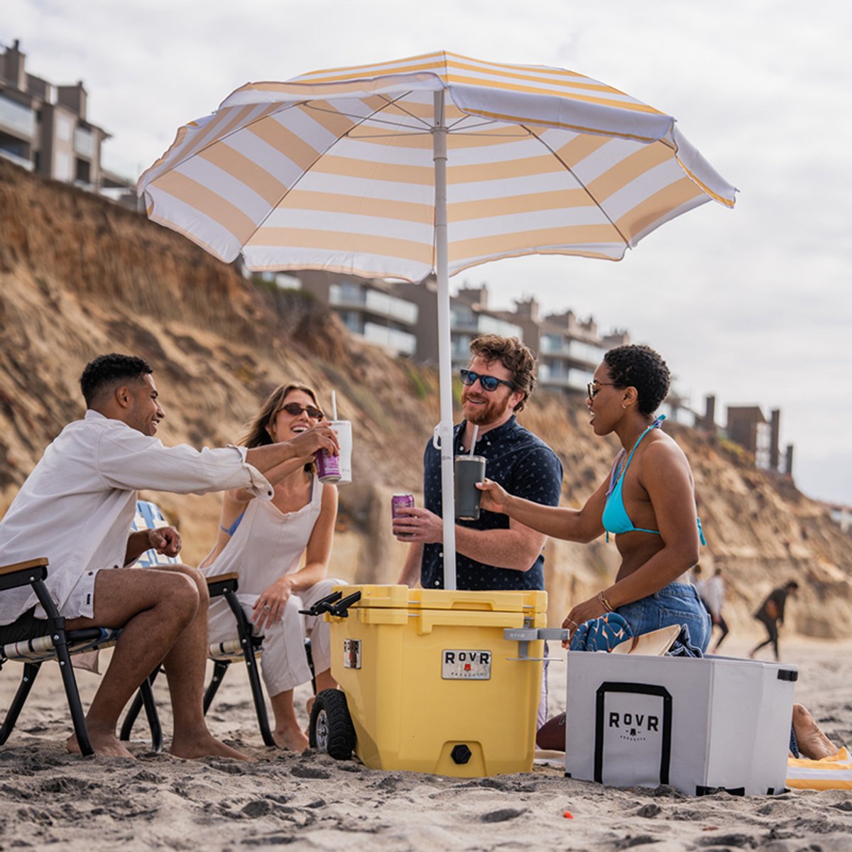 Group of friends gathered around a yellow RovR RollR cooler on the beach under a striped umbrella, raising drinks