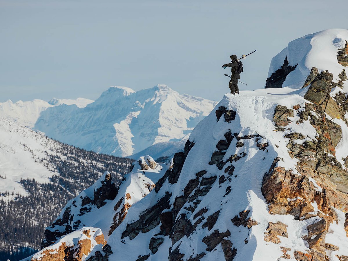 Skier wearing Backcountry Cottonwoods kit carrying skis standing on snowy peak looking out at mountain view.