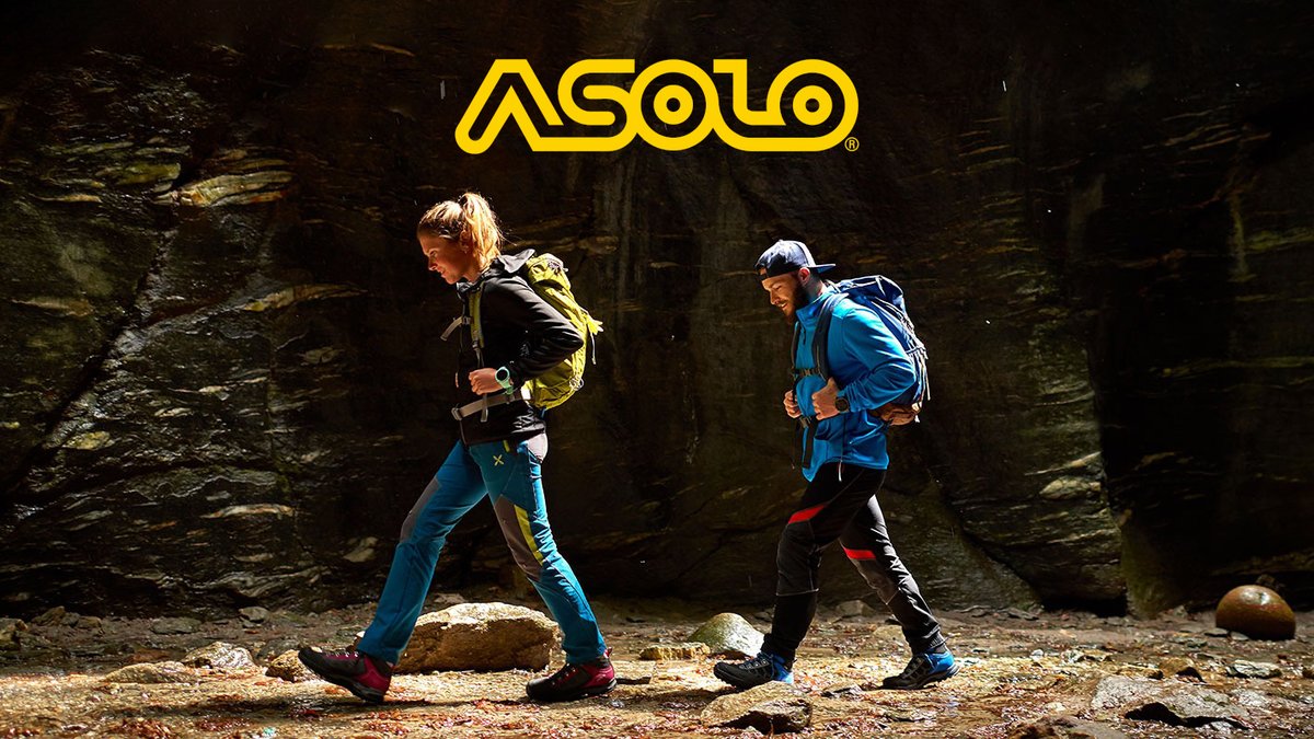 Two trail runners wearing Asolo hiking boots and backpacks run through a rocky canyon with moss-covered stone walls during an outdoor adventure.