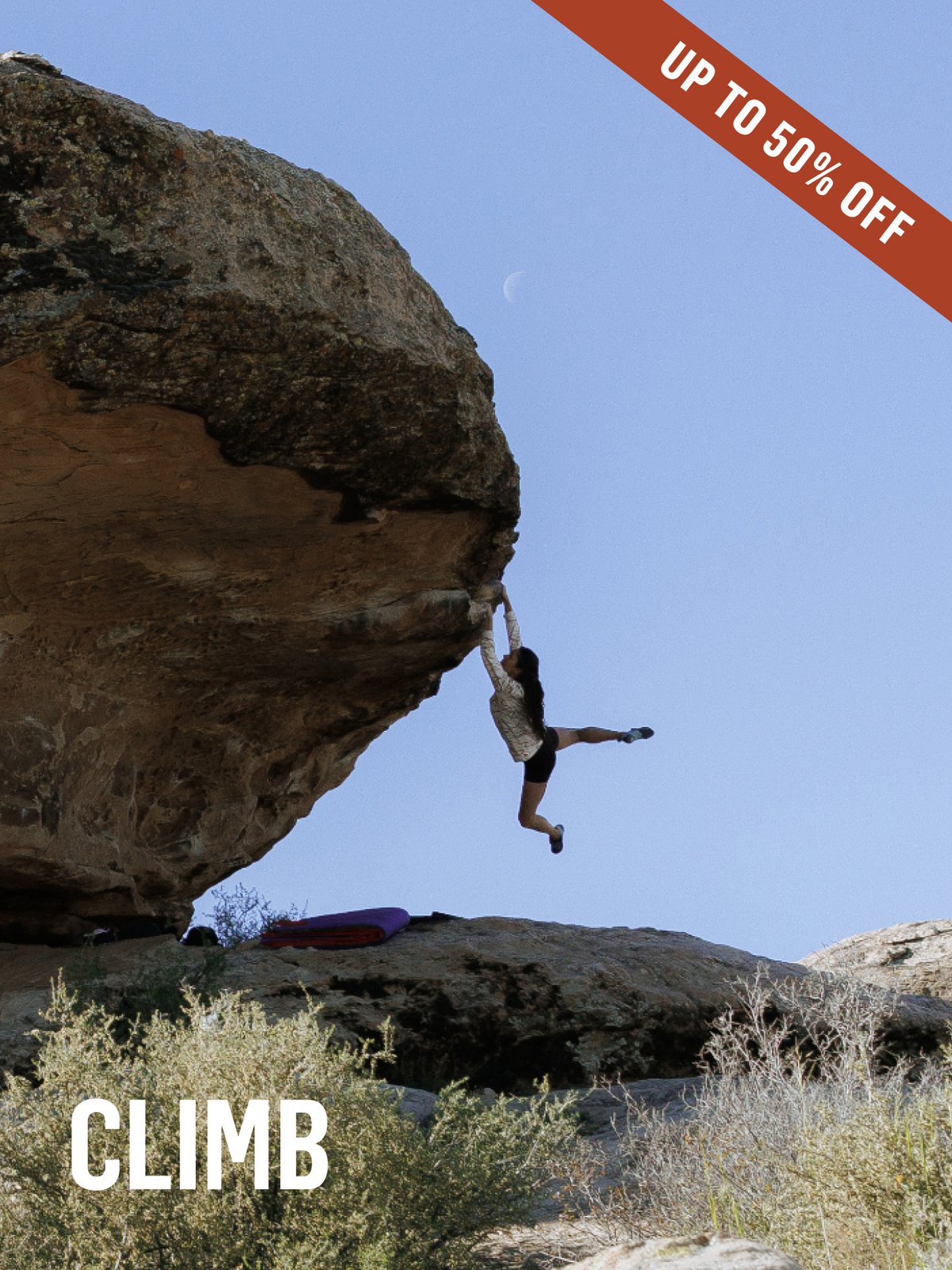 A climber hangs off a dramatic sandstone overhang, legs flagging, crash pad visible below. A crescent moon floats in the blue desert sky. Climb up to 50% off.