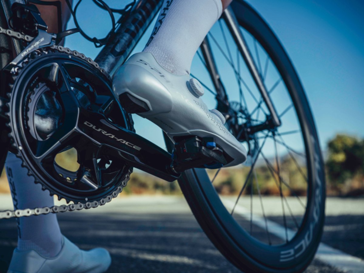 Close-up of a cyclist's foot clipped into a Shimano Dura-Ace crankset on a road bike, wearing white cycling shoes and socks.