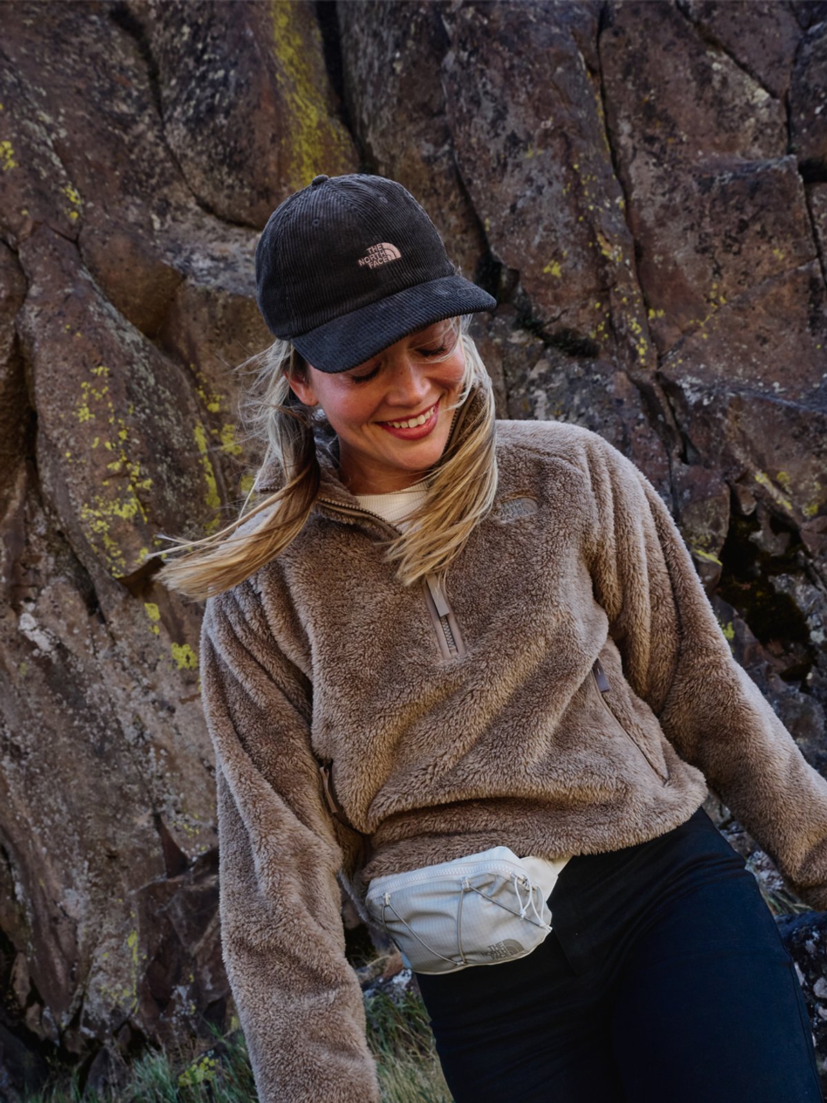 A woman wears The North Face half zip fleece, standing against a rocky background with her hair blowing in the breeze.