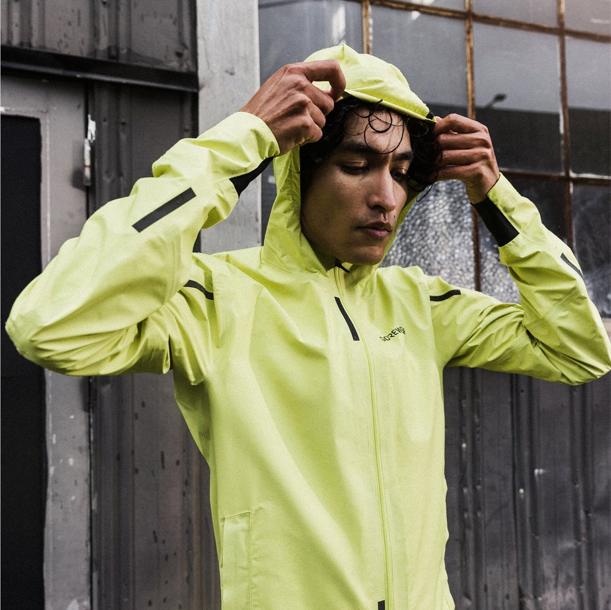 A person wearing a light yellow hooded jacket is adjusting the hood while standing in front of a metal building.