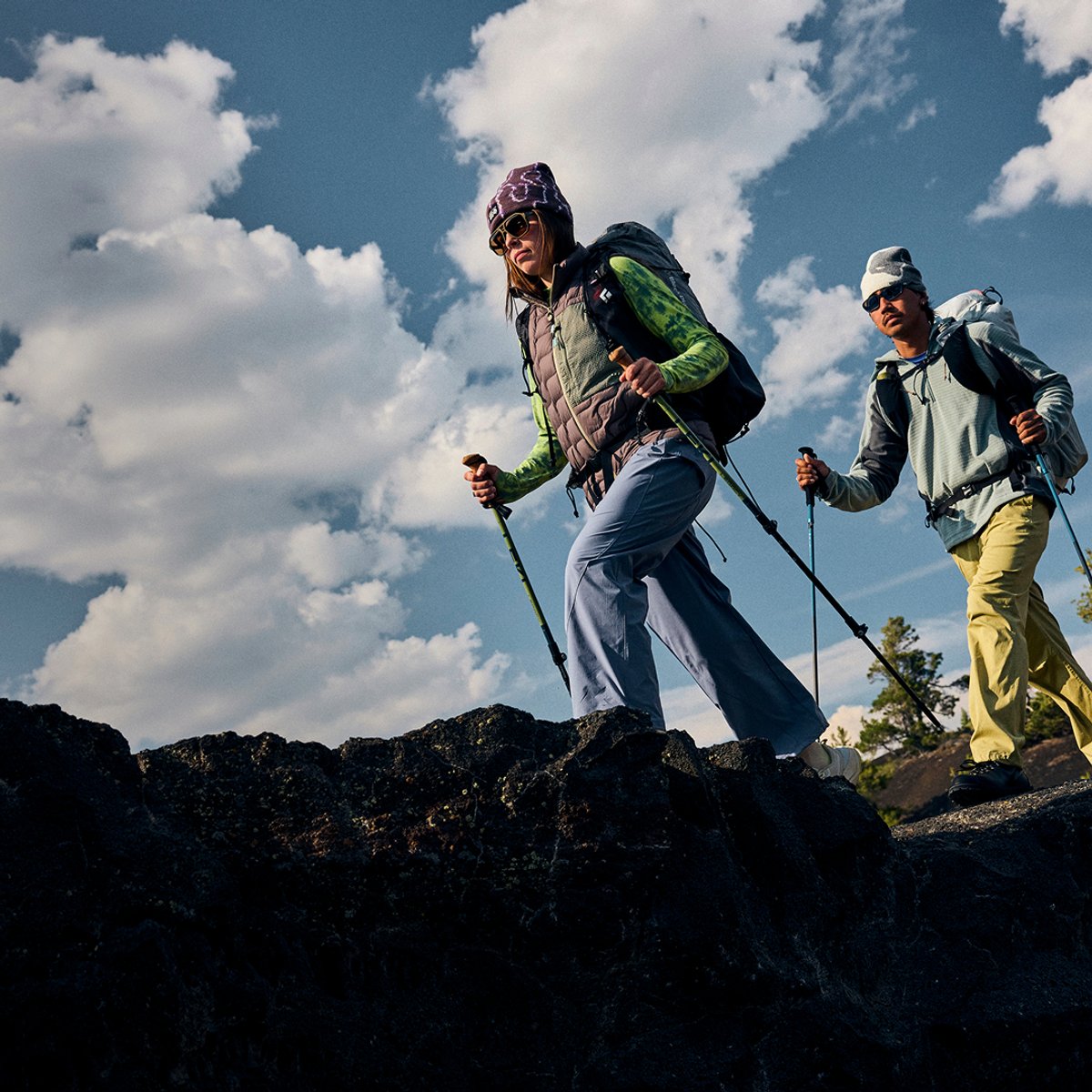 Two hikers with trekking poles and daypacks crossing a dark volcanic rock ridge under a cloudy sky