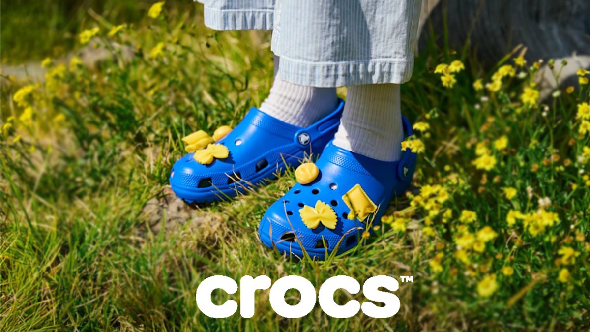 Child wearing bright blue Crocs clogs with yellow flower charms standing in a sunny grassy field with yellow wildflowers.