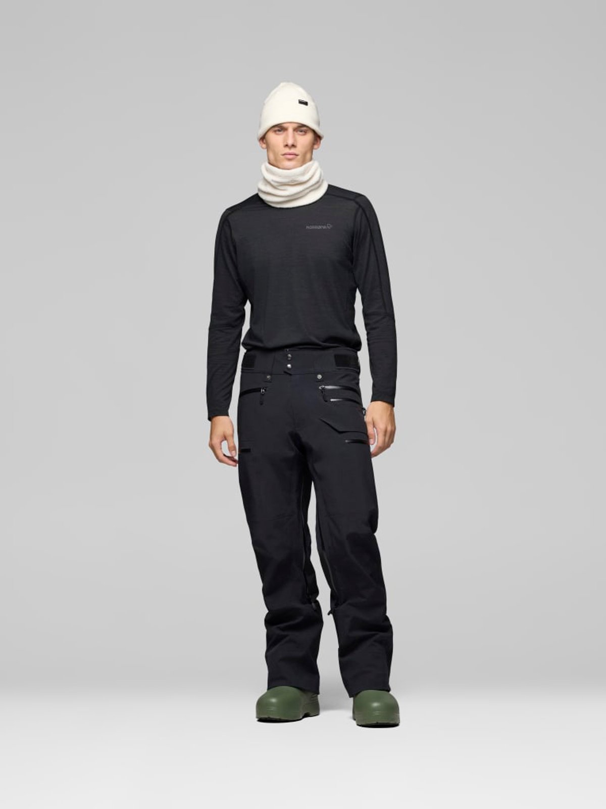 Man wearing black long-sleeve crew neck sweatshirt with white beanie and neck gaiter paired with black ski pants and green winter boots against a neutral gray background.