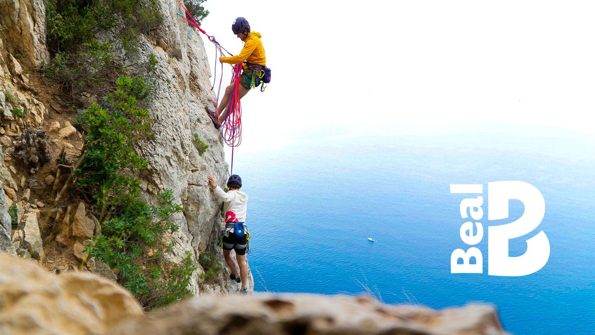 Two rock climbers rappel down a scenic coastal cliff with crystal blue ocean water in the background, demonstrating Beal climbing gear and safety equipment.