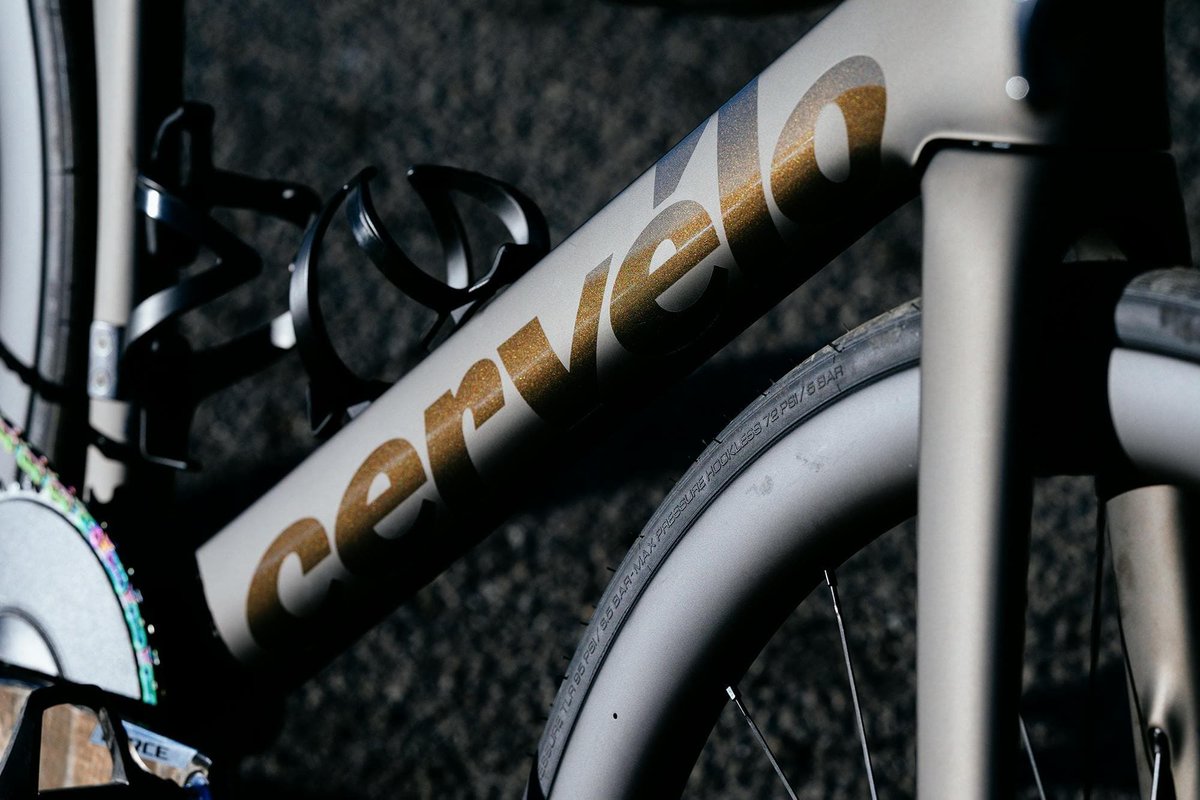 Close-up of Cervélo road bike down tube showing gold glitter logo graphics with black bottle cage and road tire detail