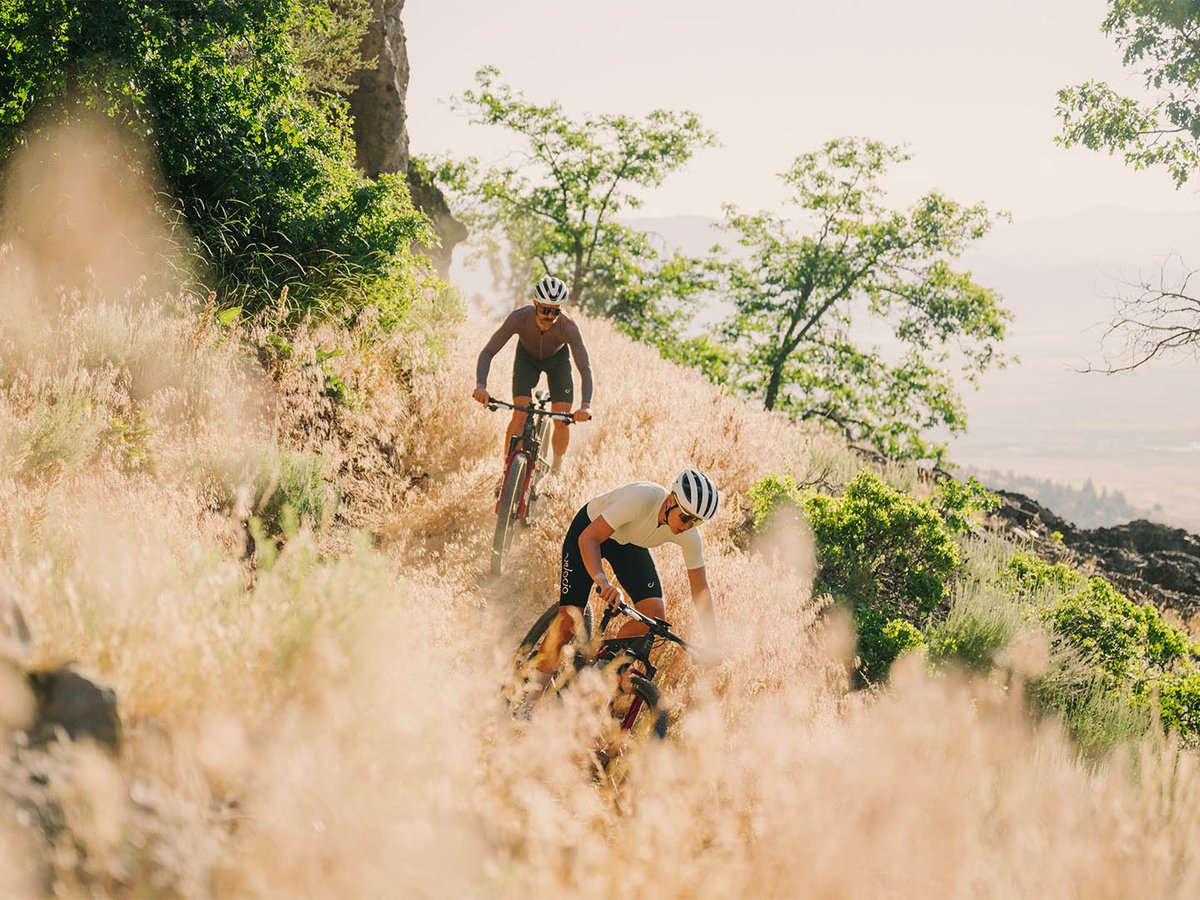Two bikers riding down a hillside trail with dry grasses all around.