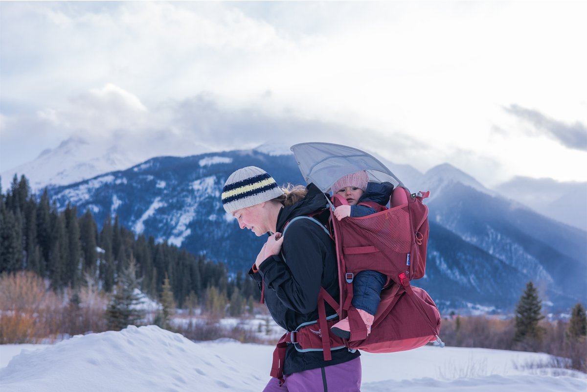 A woman hikes with a child in a red Osprey carrier with mountains behind her.