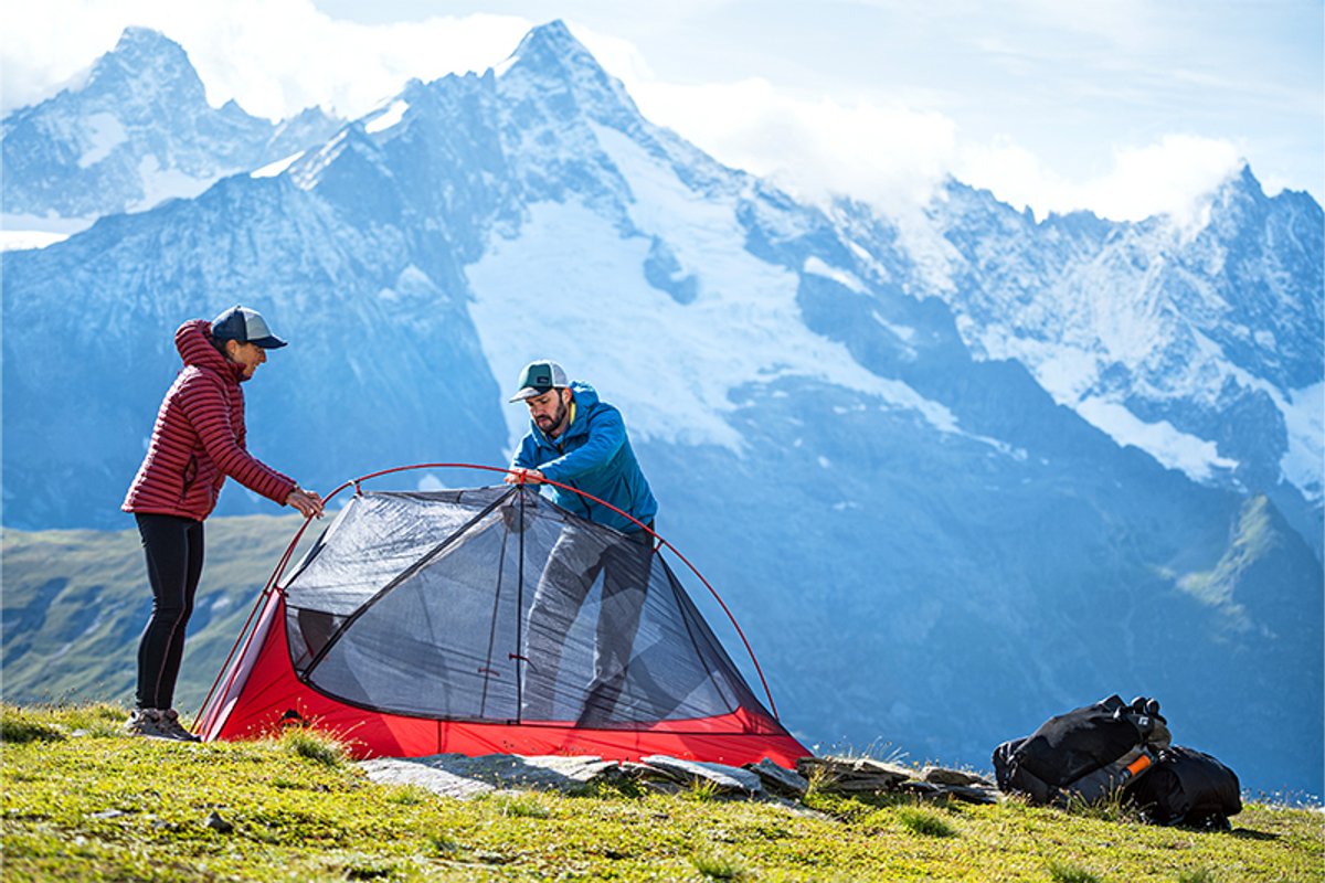 Two campers set up a tent on a grassy ridge with dramatic mountain peaks and backpacks nearby.