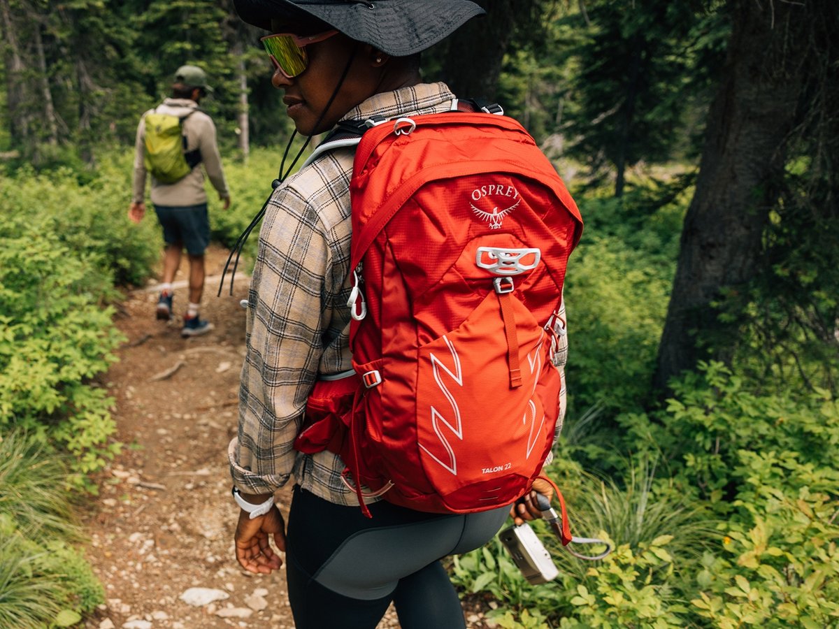 Two people wearing backpacks hike through the woods on a dirt path.