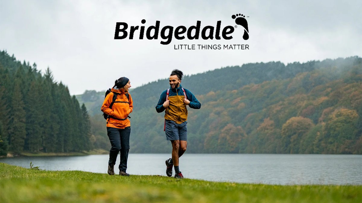 Two runners jogging along a scenic lakeside trail surrounded by forested mountains wearing colorful outdoor jacket, backpacks, and Bridgedale socks.