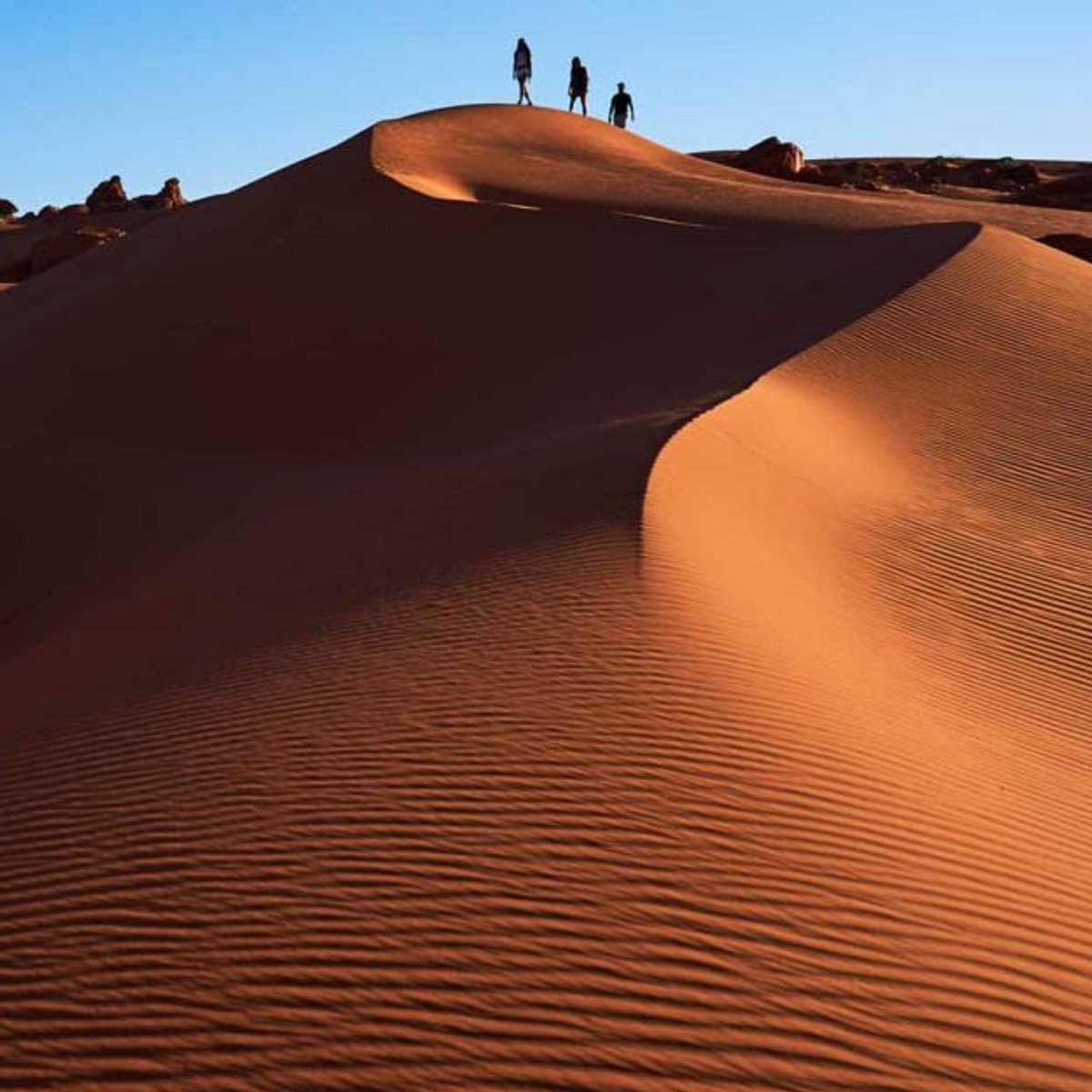 Three people silhouetted on the crest of a large red sand dune at Sand Hollow, with eroded sandstone outcrops along the left edge and a clear blue sky above.