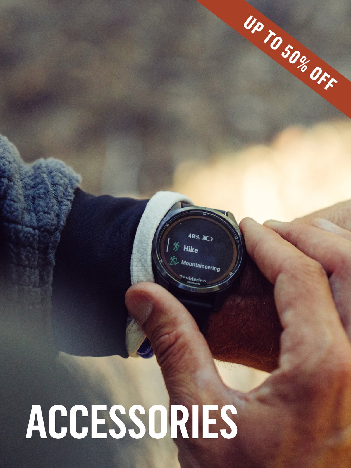 A Garmin GPS watch displaying Hike and Mountaineering activity modes, being navigated by a gloved hand on a rocky outcrop. Up to 50% off accessories.