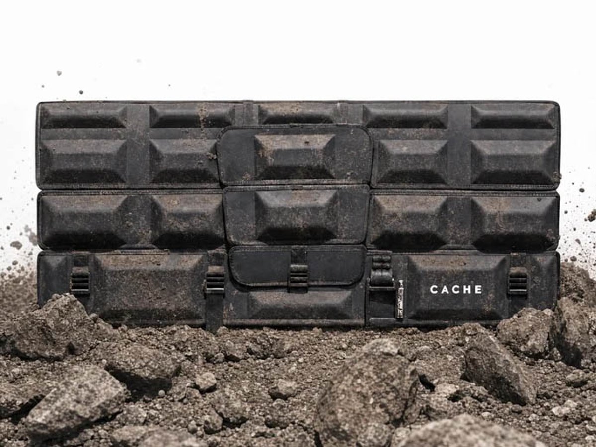 A Cache Basecamp tailgate pad standing upright in a pile of dirt and rock against a white background, with the CACHE wordmark at the lower right.