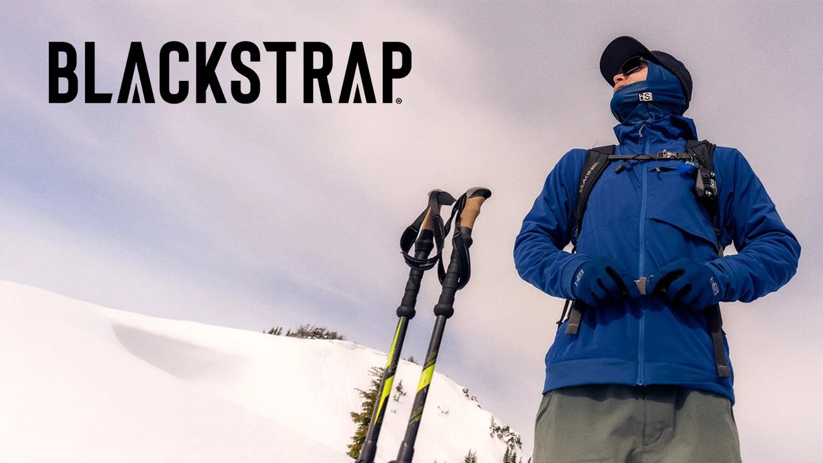 A person in a blue technical jacket and face mask stands in a snowy landscape next to a pair of ski poles.
