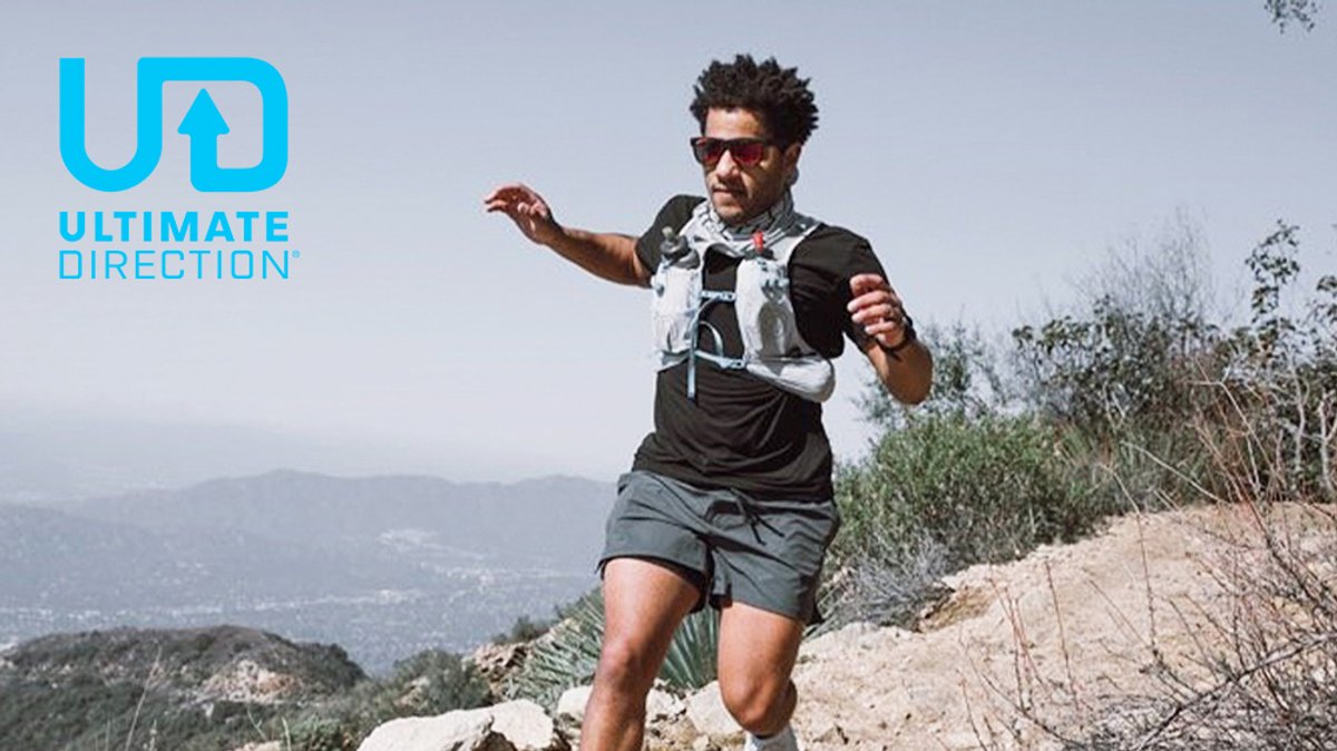 Man trail running downhill on a mountain path wearing a black and gray athletic outfit with a Ultimate Direction hydration pack and sunglasses against a scenic valley landscape.