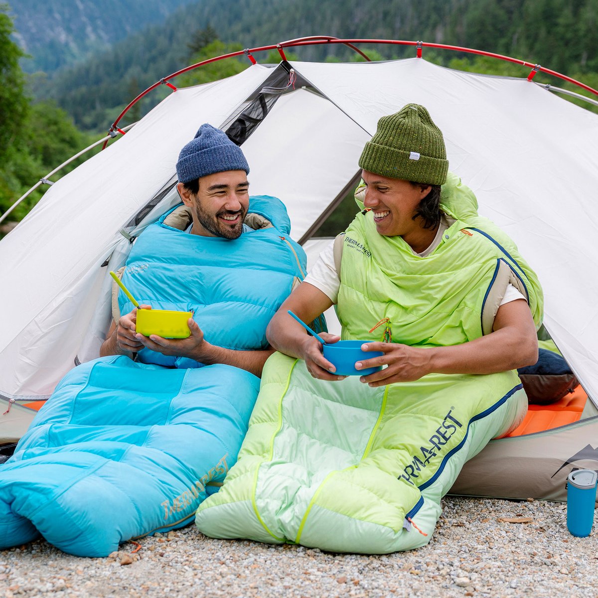 Two backpackers stay warm cocooned in their sleeping bags while eating breakfast. Their sleeping bags have arm-hole openings to allow for freedom of movement.