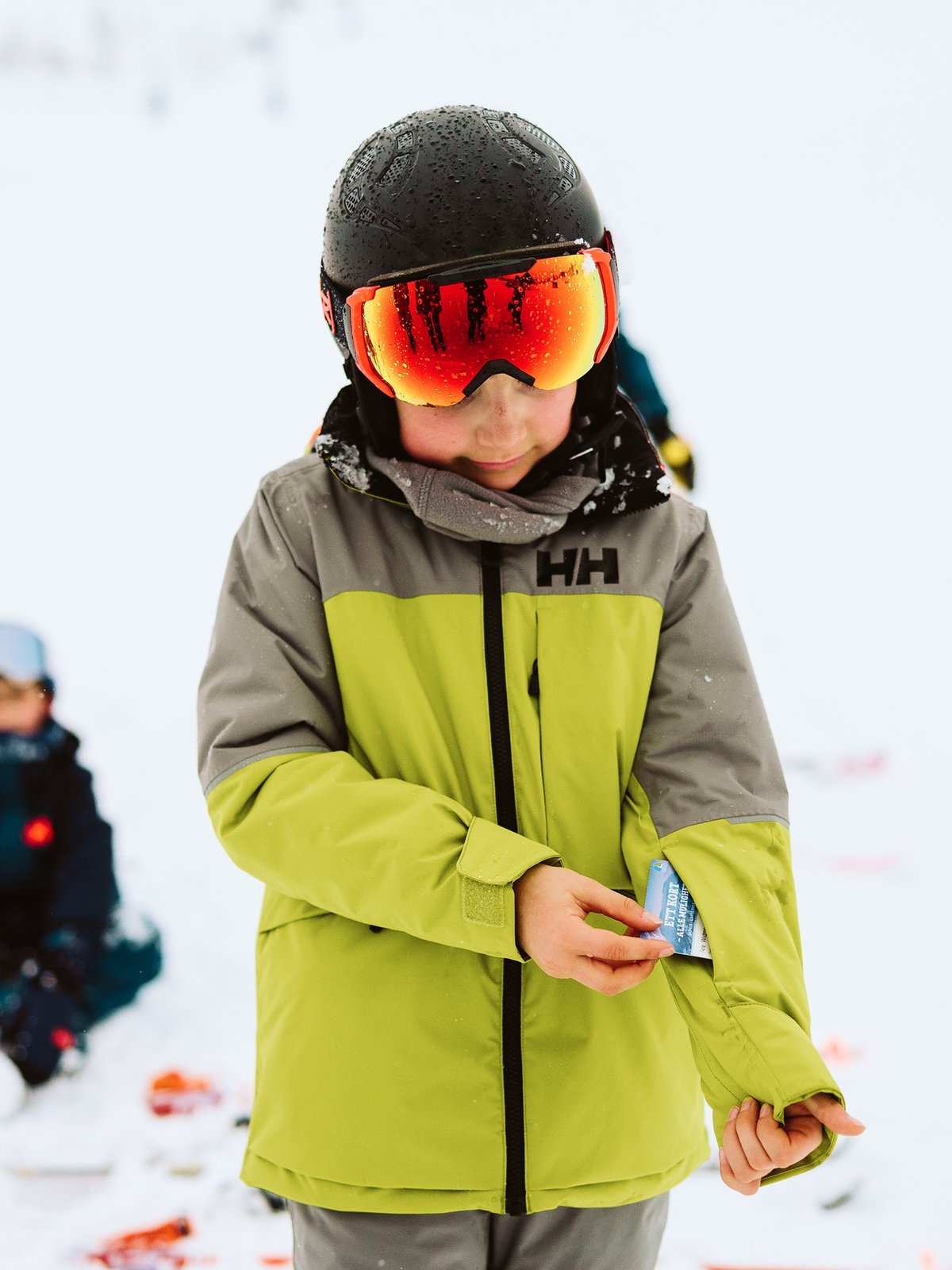 A child wearing a lime green and gray Helly Hansen jacket, black helmet, and orange ski goggles stands in the snow, with another child sitting in the snowy background.