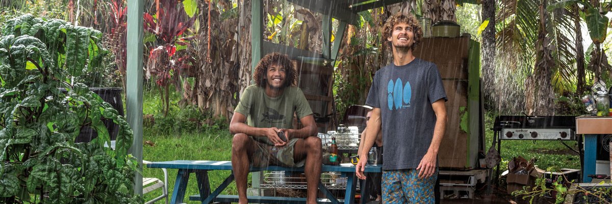 Two men enjoy a covered outdoor space during heavy rain, surrounded by lush tropical vegetation and camping gear, showcasing weather-resistant shelter solutions for outdoor adventures.