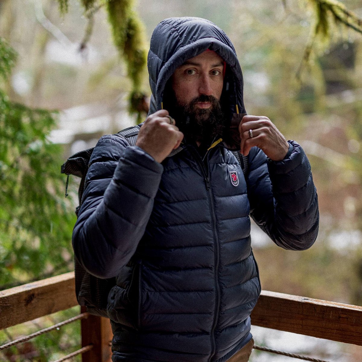 A man wears a Fjallraven Expedition jacket with the hood up, standing against a forest background.