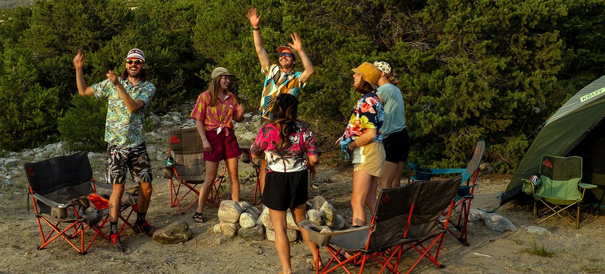 A group of friends stand around a fire pit, smiling and laughing. The Kelty Loveseat is featured prominently behind the group.