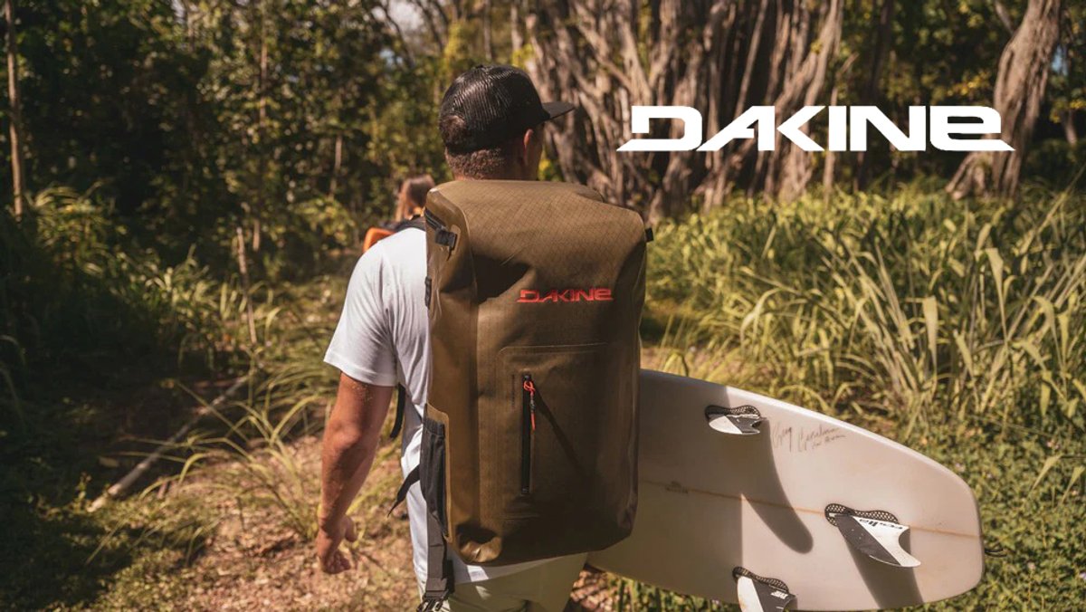 A man walks through tropical greenery carrying a surfboard and wearing a large, olive-green Dakine waterproof backpack.