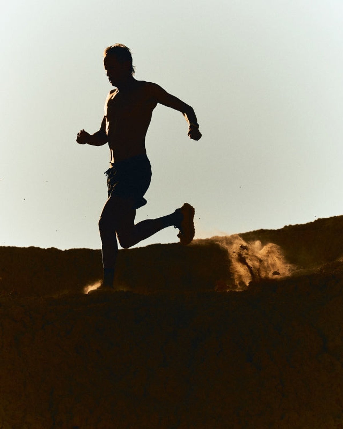 Silhouette of a shirtless male runner in mid-stride along a dusty ridgeline, backlit against a pale sky with dust kicking up behind.