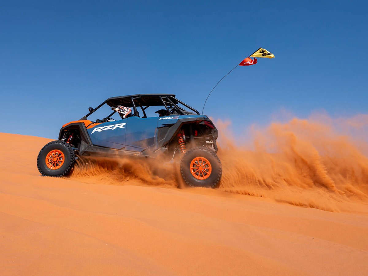 Blue and orange Polaris RZR Pro side-by-side UTV kicking up sand while turning on a red sand dune under a clear blue sky.