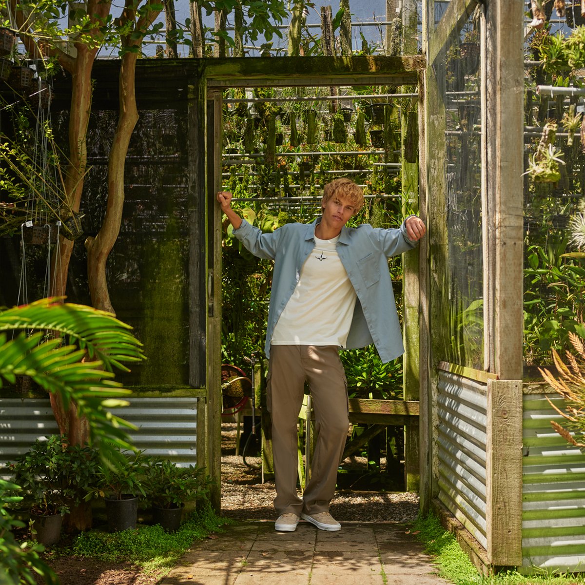 A man wears the prAna Zion Stretch Pants, leaning against the doorframe of a greenhouse.
