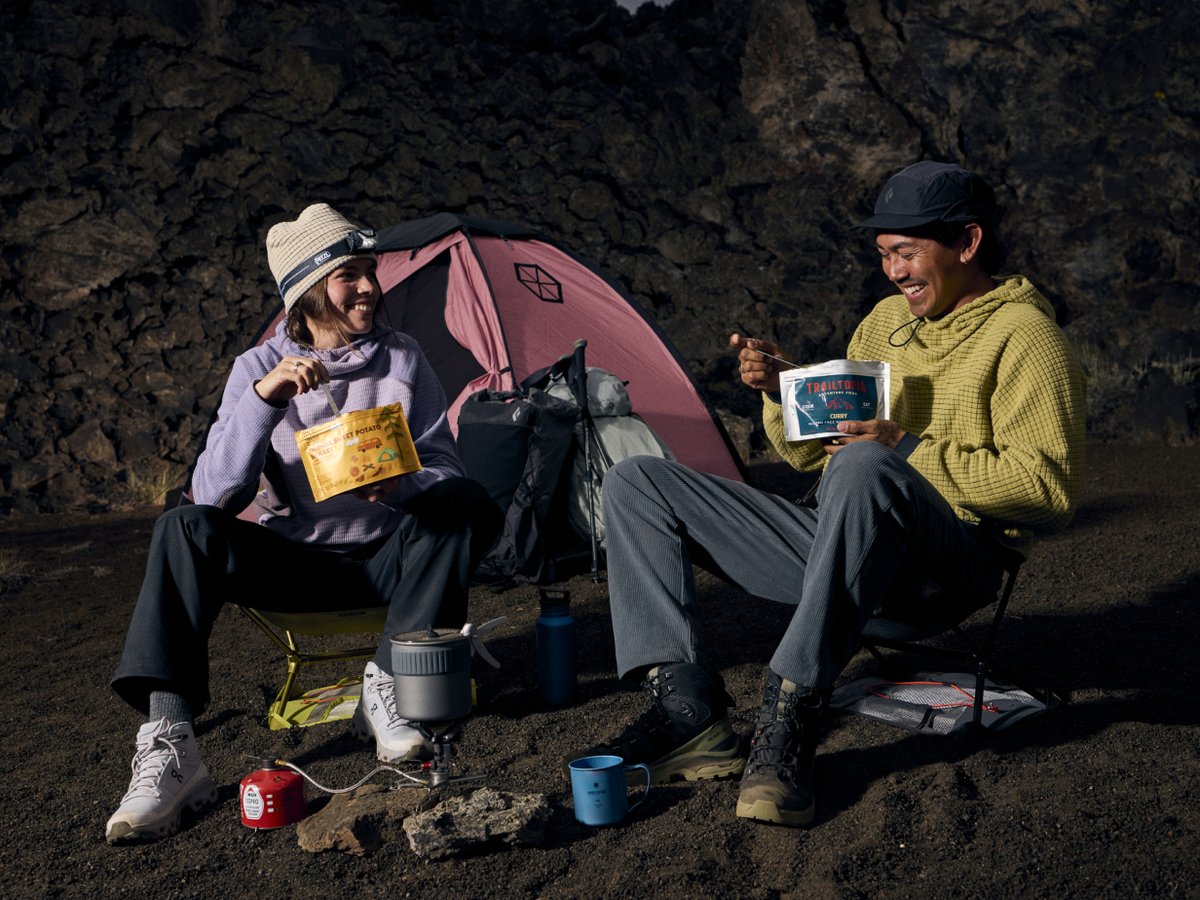 Two campers eating freeze-dried meals on volcanic terrain, laughing beside a pink tent and camp stove at dusk.