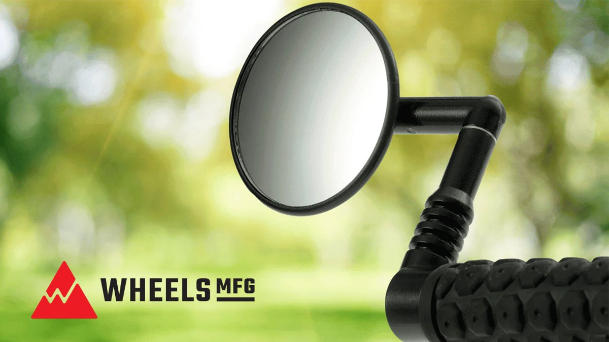 Black bicycle handlebar mirror with ergonomic grip and wide viewing angle against blurred green outdoor background from Wheels MFG.
