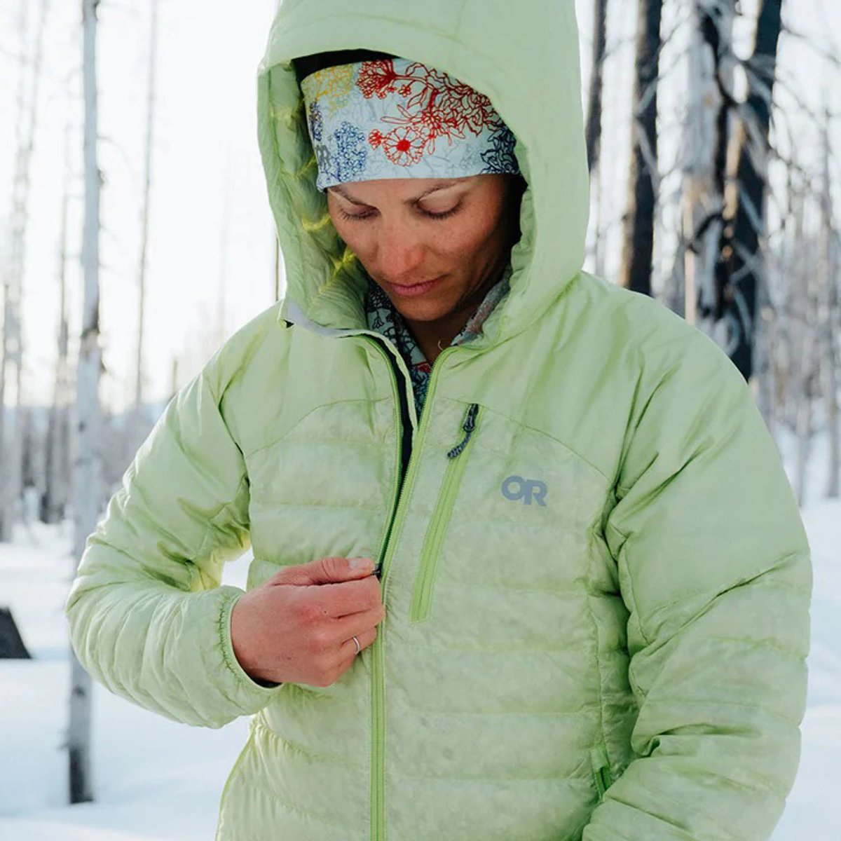 A woman in a snowy forest zips up the Helium Down Hoodie. 