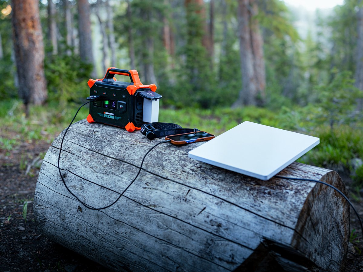 A Dark Energy Recon power station sits on a fallen log in the woods charging a phone, with a headlamp and a laptop beside it.