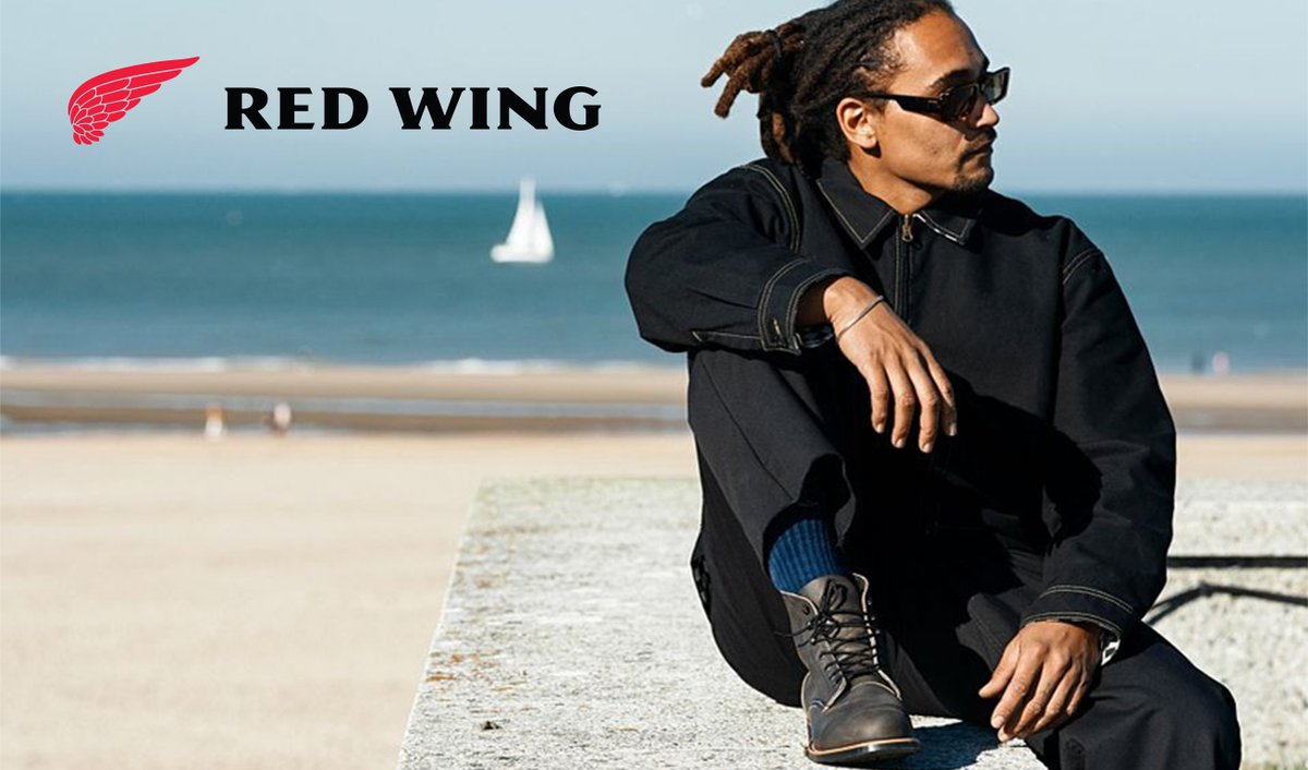 A man wearing black Red Wing boots and casual apparel sits on a beach jetty with the ocean and sailboat visible in the background.